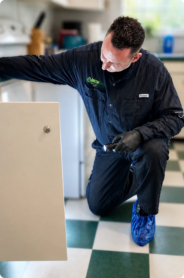 A technician in a dark uniform kneels on a checkered kitchen floor inspecting a cabinet panel