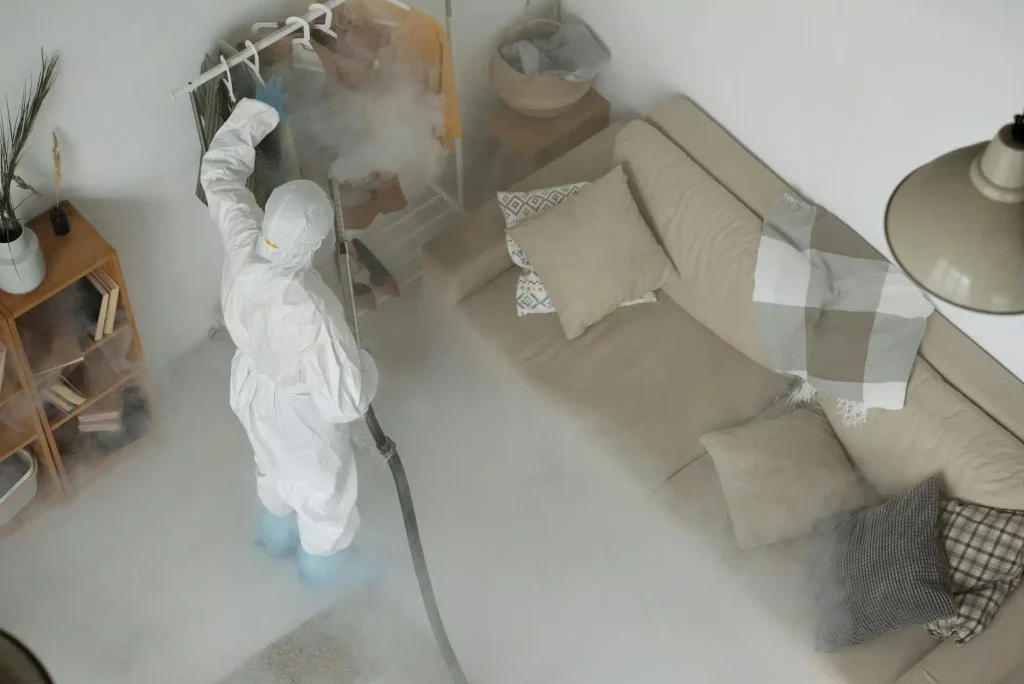 Person in white hazmat suit spraying disinfectant fog throughout a living room with a beige sofa
