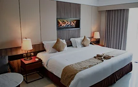 Neatly made king-size hotel bed with white linens, gold accent runner, flanking nightstands with lamps, and wall art above headboard