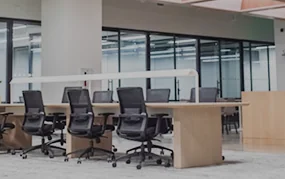 Open-plan office with rows of wooden desks and black mesh ergonomic chairs in a modern, well-lit workspace